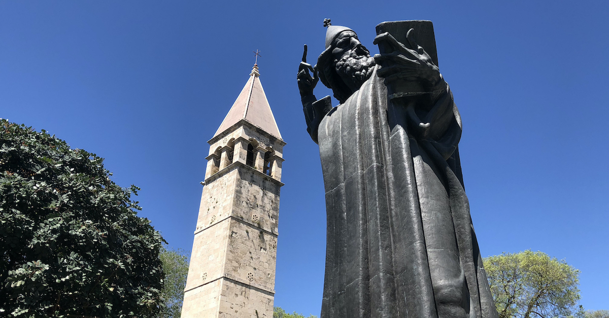 Tall statue of Gregory of Nin reading from a document with a bell tower looming in the background