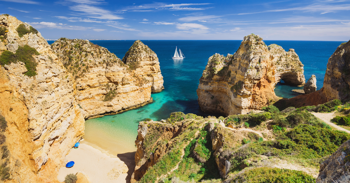 Rocky golden coastline leading into deep blue ocean with a white sailboat in the distance