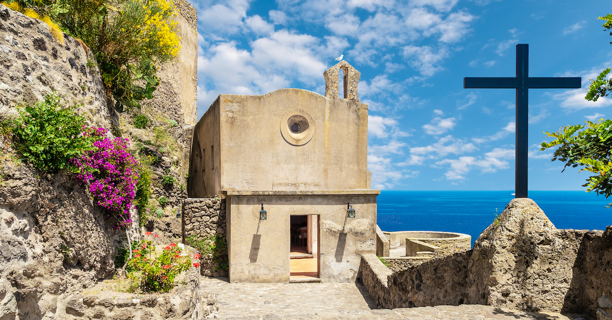 Small beige stone church with a tall black cross next to it and ocean in the background