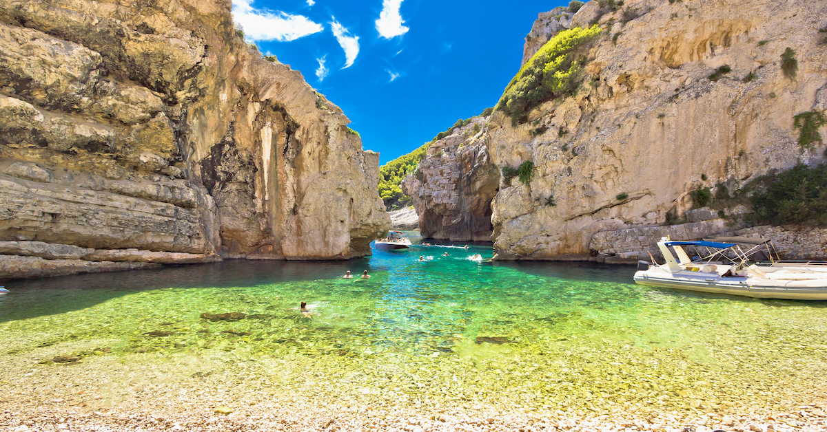 Clear water on and rocky shore in the foreground with two small boats on the right leading out an inlet between two beige cliff in Stiniva Bay on Vis island, one of the unique Croatia honeymoon destinations