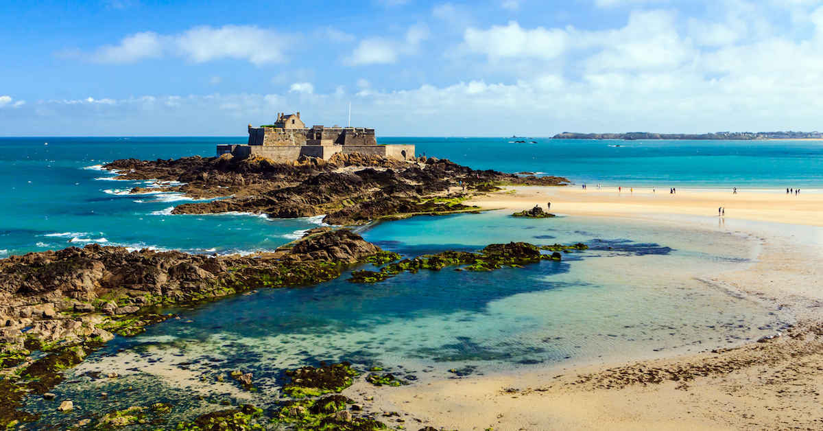 Brown rocks in blue water just off of a sandy coast leading up to a stone building in Saint Malo, France
