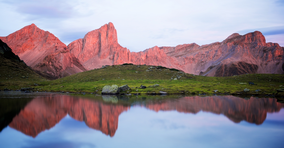 Background mountains tinted pink by sunset reflected in a lake in the foreground with green hills rolling through the middle ground