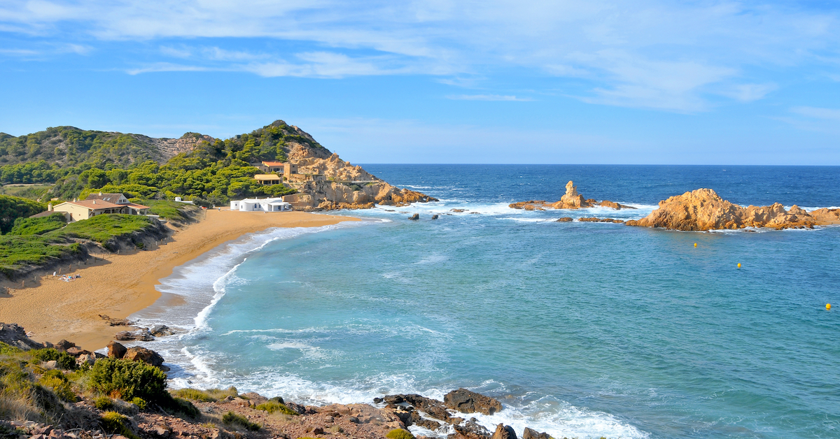 Green hills and golden sand beach on the right leading to blue ocean on the left in Menorca of the Balearic Islands, one of many ideal Spain honeymoon destinations