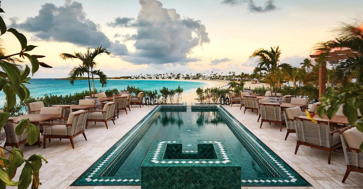 Narrow pool lined with table in chairs in the foreground leading towards a sandy coast wrapping around blue water with white buildings in the distance 