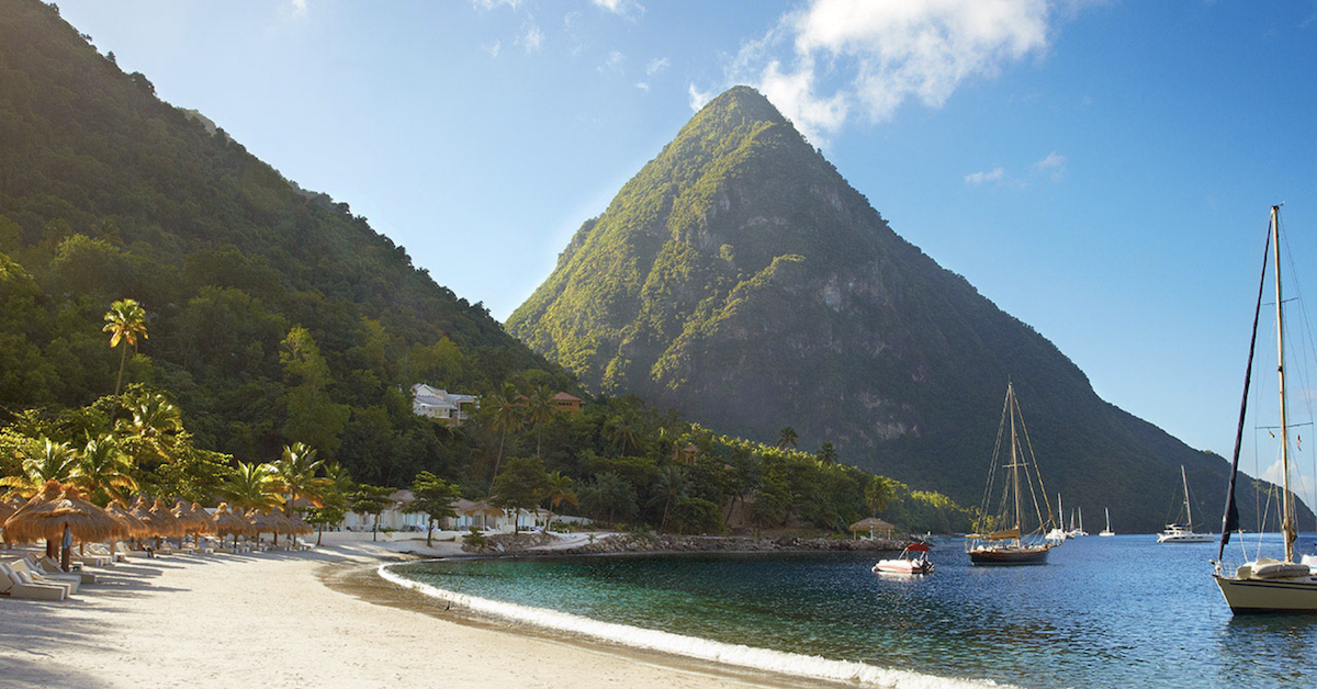 Sandy beach lined with umbrellas and lounge chairs and several boats in the water with a green mountain in the background