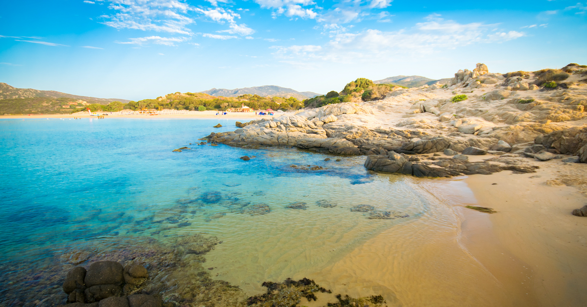 Chia Beach in Sardinia, Italy, made of golden sands and clear blue waters with trees and mountains in the distance