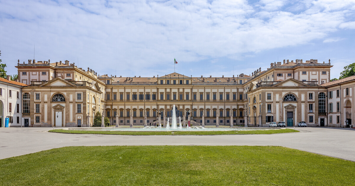 A sprawling tan and white palace with a fountain in front and an Italian flag on top